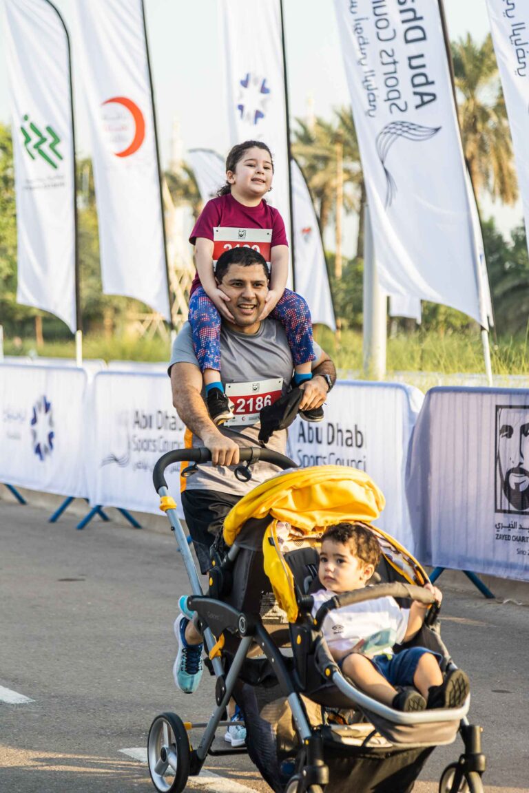 Man with child on top of shoulders running with a stroller in the Zayed Charity Marathon Abu Dhabi 2022